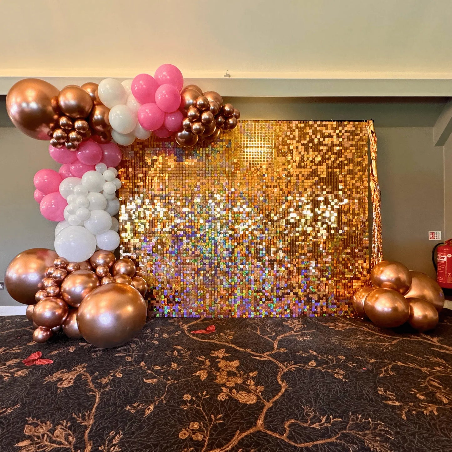 A luxurious event setup featuring a shimmering rose gold sequin wall adorned with a premium balloon garland. The garland includes an elegant mix of rose gold, pink, and white balloons in varying sizes, creating a stunning backdrop perfect for weddings, parties, or special occasions. | The Party Hut Cheltenham